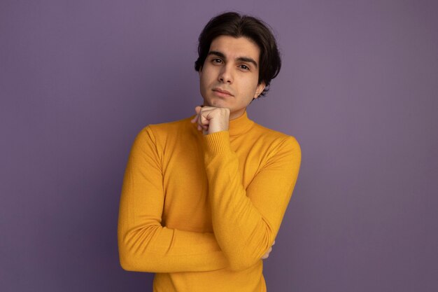 Fiducioso giovane bel ragazzo che indossa un maglione dolcevita giallo mettendo la mano sotto il mento isolato sulla parete viola