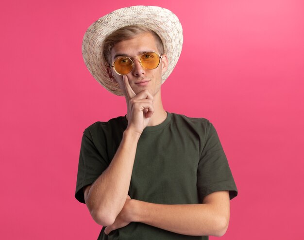 Fiducioso giovane bel ragazzo indossa camicia verde e occhiali con il cappello che mette il dito sulla guancia isolato sul muro rosa