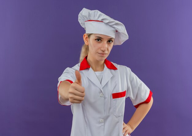 Fiducioso giovane bella cuoca in uniforme da chef mettendo la mano sulla vita e mostrando il pollice in alto isolato sulla parete viola con spazio copia