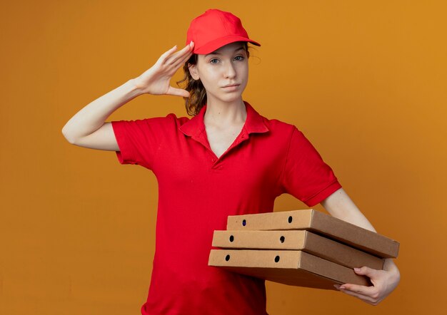 Fiducioso giovane bella ragazza di consegna in uniforme rossa e cappuccio tenendo i pacchetti di pizza e facendo gesto di saluto isolato su sfondo arancione