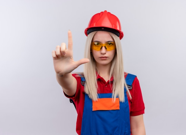 Fiducioso giovane bionda ingegnere builder ragazza in uniforme con gli occhiali di sicurezza che mostra una pistola simile a una pistola su uno spazio bianco isolato con spazio di copia