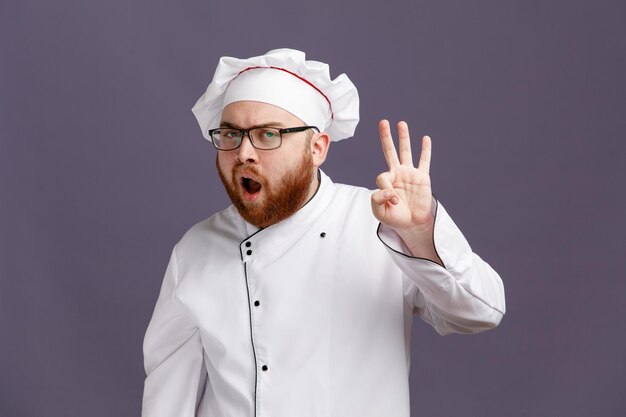 Fiducioso giovane chef indossando occhiali uniforme e cappuccio guardando la fotocamera che mostra segno ok isolato su sfondo viola