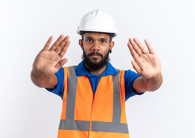Fiducioso giovane costruttore afro-americano uomo in uniforme con casco di sicurezza che gesturing il segnale di stop con due mani isolate su sfondo bianco con spazio di copia