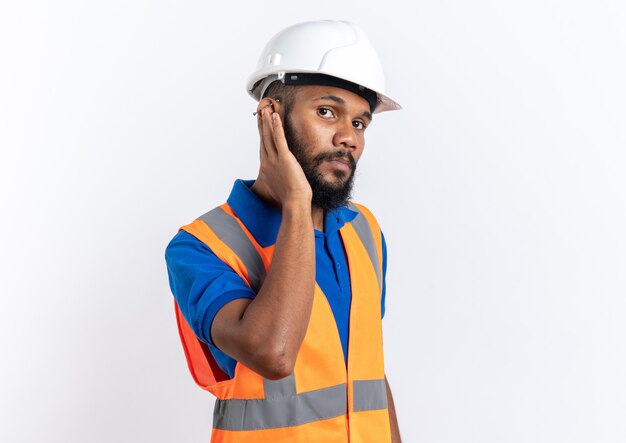 Fiducioso giovane costruttore afro-americano uomo in uniforme con casco di sicurezza mettendo la mano sull'orecchio isolato su sfondo bianco con spazio di copia