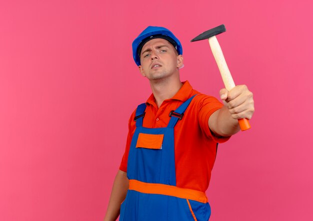 Fiducioso giovane costruttore maschio indossa uniforme e casco di sicurezza porgendo un martello