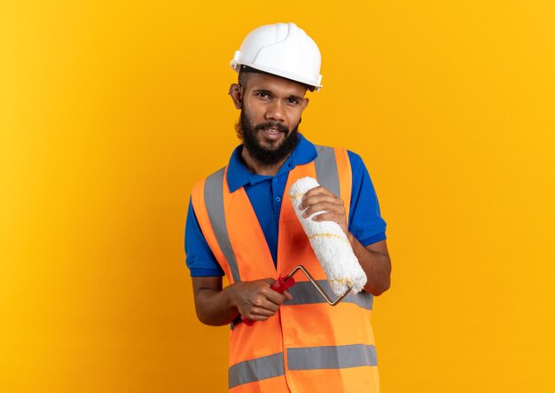 fiducioso giovane costruttore uomo in uniforme con casco di sicurezza tenendo il rullo di vernice isolato sulla parete arancione con copia spazio
