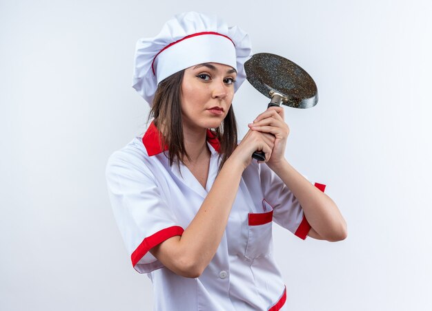 Fiducioso giovane cuoca che indossa l'uniforme dello chef tenendo la padella isolata su sfondo bianco