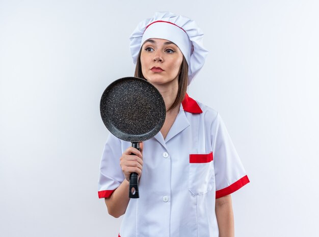 Fiducioso giovane cuoca che indossa l'uniforme dello chef tenendo la padella isolata sul muro bianco