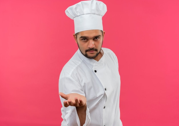 Fiducioso giovane cuoco maschio in uniforme da chef che allunga la mano isolata sulla parete rosa con spazio di copia