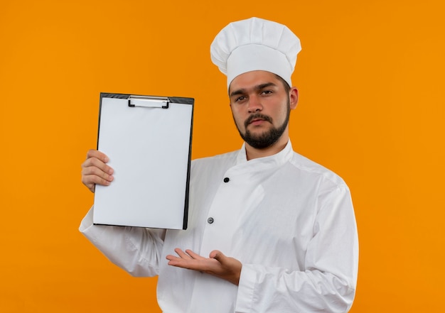 Fiducioso giovane cuoco maschio in uniforme da chef che mostra e indica con la mano la lavagna per appunti isolata sulla parete arancione