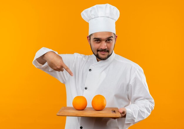 Fiducioso giovane cuoco maschio in uniforme da chef che tiene e punta al tagliere con arance su di esso isolato su parete arancione