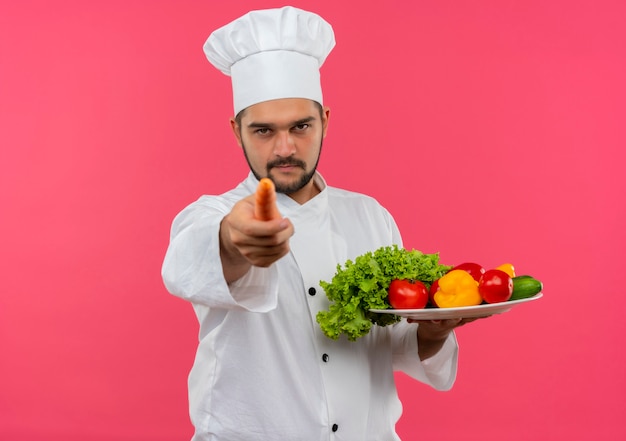 Fiducioso giovane cuoco maschio in uniforme da chef che tiene piatto di verdure e indica con la carota isolata sulla parete rosa con spazio di copia