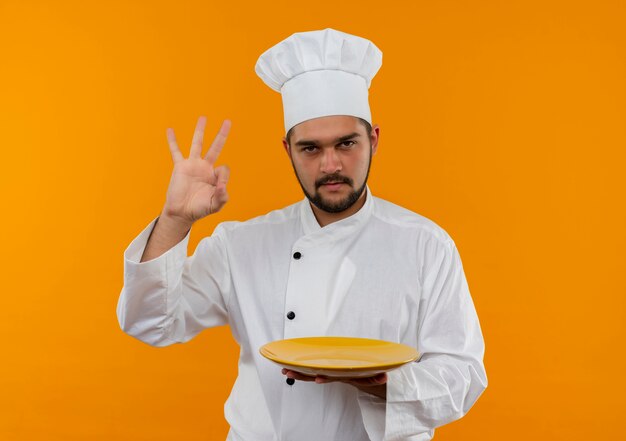 Fiducioso giovane cuoco maschio in uniforme da chef che tiene piatto vuoto e fa segno ok isolato sulla parete arancione con spazio copia