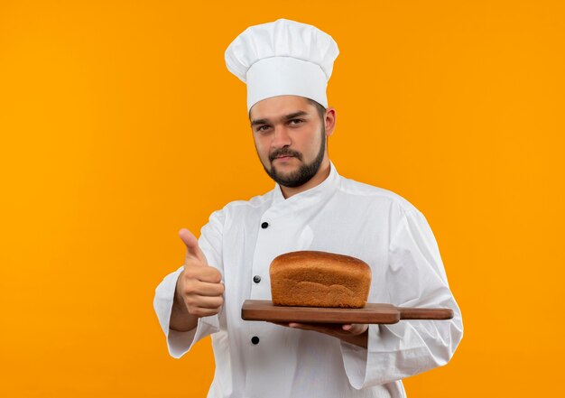 Fiducioso giovane cuoco maschio in uniforme da chef che tiene tagliere con pane su di esso che mostra pollice in su isolato su parete arancione orange