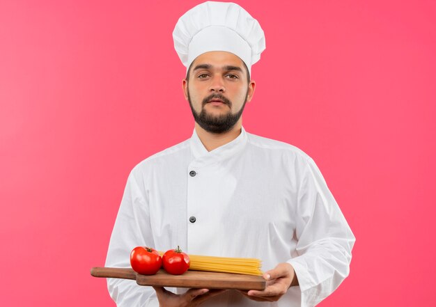 Fiducioso giovane cuoco maschio in uniforme da chef che tiene tagliere con pomodori e pasta di spaghetti su di esso isolato su parete rosa