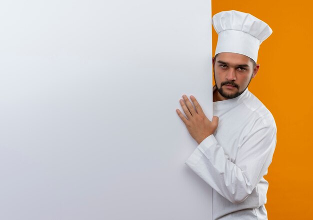 Fiducioso giovane cuoco maschio in uniforme da chef in piedi dietro e mettendo la mano sul muro bianco isolato sul muro arancione con spazio copia