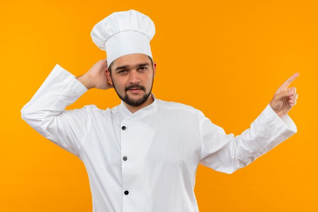 Fiducioso giovane cuoco maschio in uniforme da chef mettendo la mano dietro la testa e indicando il lato isolato sulla parete arancione