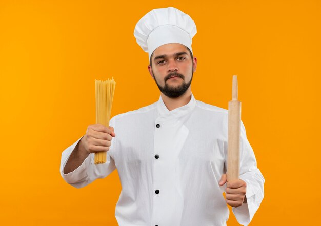 Fiducioso giovane cuoco maschio in uniforme da chef tenendo gli spaghetti e mattarello isolato su parete arancione