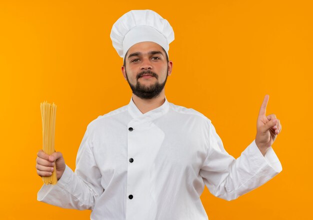 Fiducioso giovane cuoco maschio in uniforme da chef tenendo gli spaghetti e rivolto verso l'alto isolato su parete arancione