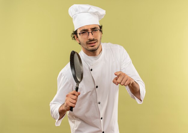 fiducioso giovane cuoco maschio indossa uniforme da chef e bicchieri tenendo padella e mostrandoti gesto