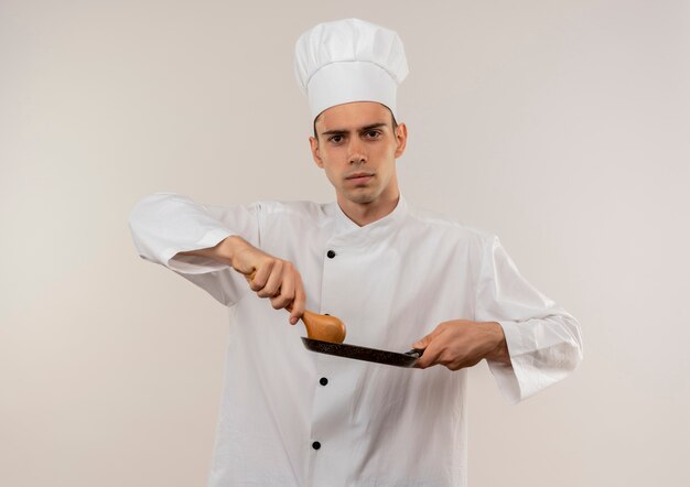 Fiducioso giovane cuoco maschio indossa uniforme del cuoco unico che tiene padella e cucchiaio sulla parete bianca isolata
