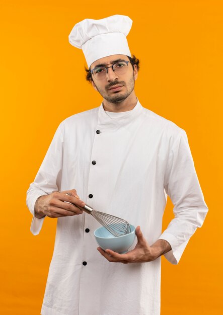 Fiducioso giovane cuoco maschio indossando l'uniforme del cuoco unico e vetri che tengono frusta e ciotola