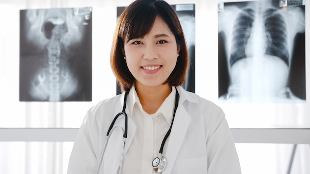 Fiducioso giovane dottoressa asiatica in uniforme medica bianca con stetoscopio che guarda l'obbiettivo e sorride durante la videoconferenza con il paziente in ospedale.