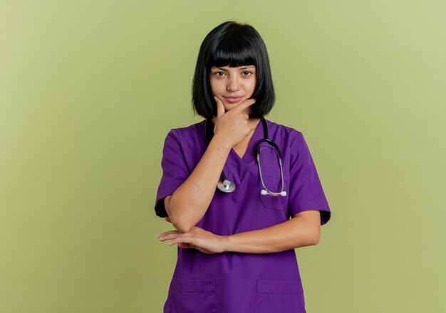 Fiducioso giovane dottoressa bruna in uniforme con lo stetoscopio mette la mano sul mento isolato su sfondo verde oliva con lo spazio della copia