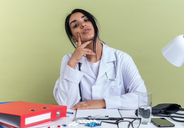 Fiducioso giovane dottoressa che indossa abito medico con stetoscopio si siede alla scrivania con strumenti medici mettendo la mano sulla guancia isolata sulla parete verde oliva