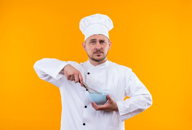 Fiducioso giovane e bello cuoco in uniforme da chef che tiene in mano una frusta e una ciotola isolate sulla parete arancione