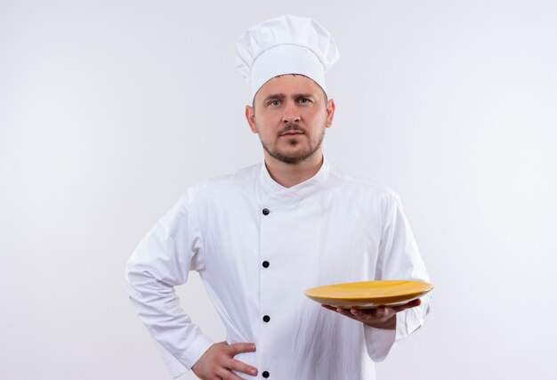 Fiducioso giovane e bello cuoco in uniforme da chef che tiene piatto con un'altra mano sulla vita sul muro bianco isolato