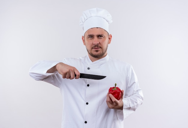 Fiducioso giovane e bello cuoco in uniforme da chef tenendo pepe e puntando con il coltello contro di esso isolato su muro bianco