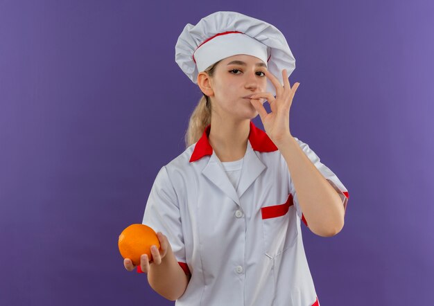 Fiducioso giovane e grazioso cuoco in uniforme da chef che fa un gesto gustoso e tiene in mano un'arancia che sembra isolata sul muro viola