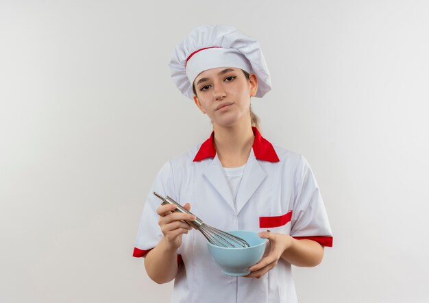 Fiducioso giovane e grazioso cuoco in uniforme da chef che tiene in mano una frusta e una ciotola isolate su un muro bianco con spazio per le copie