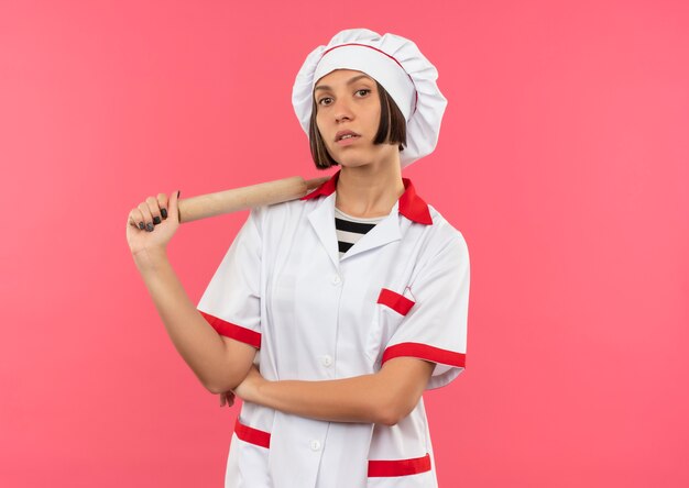 Fiducioso giovane femmina cuoco in uniforme del cuoco unico che tiene il mattarello che sembra isolato sul rosa con lo spazio della copia
