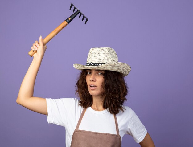 Fiducioso giovane giardiniere femminile in uniforme che indossa cappello da giardinaggio innalzamento del rastrello