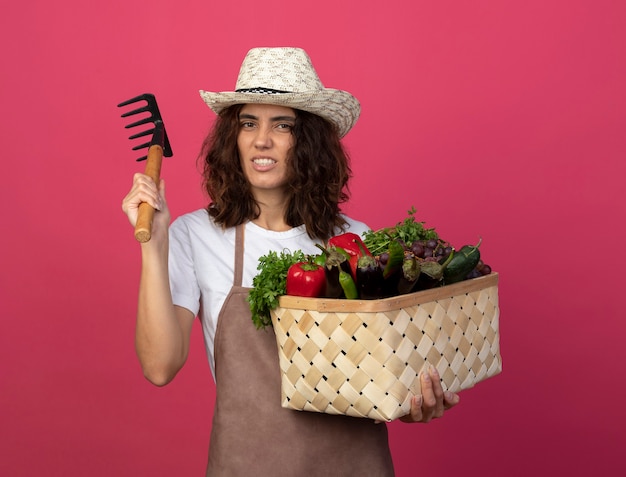 Fiducioso giovane giardiniere femminile in uniforme che indossa cappello da giardinaggio tenendo il cesto di verdure con rastrello