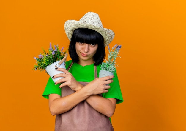 Fiducioso giovane giardiniere femminile in uniforme che indossa il cappello da giardinaggio tiene vasi di fiori che attraversano le mani