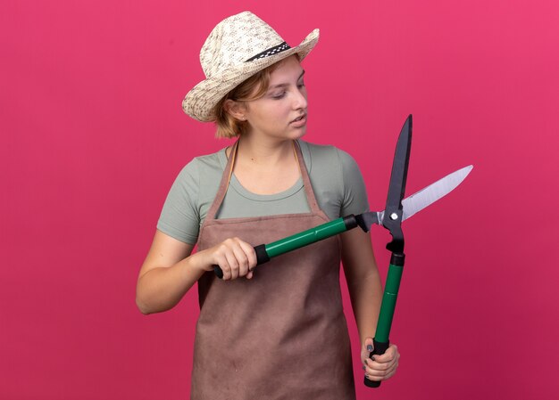 Fiducioso giovane giardiniere femminile slavo che indossa cappello da giardinaggio tenendo e guardando le forbici da giardinaggio sul rosa