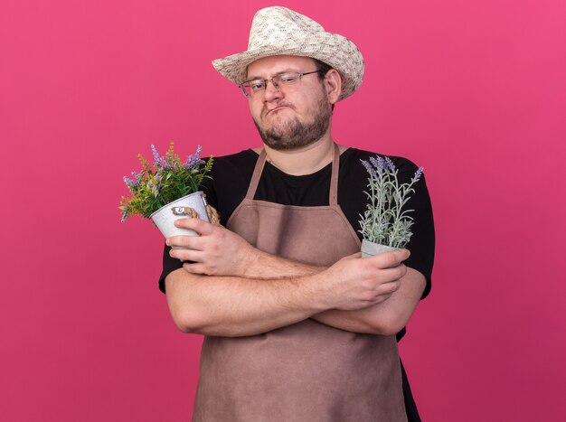 Fiducioso giovane giardiniere maschio che indossa cappello da giardinaggio tenendo e incrocio di fiori in vasi da fiori isolati sulla parete rosa