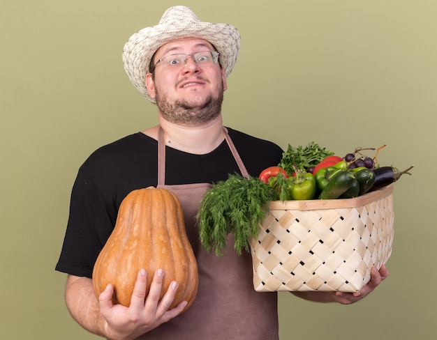 Fiducioso giovane giardiniere maschio che indossa cappello da giardinaggio tenendo il cesto di verdure con zucca isolato sulla parete verde oliva