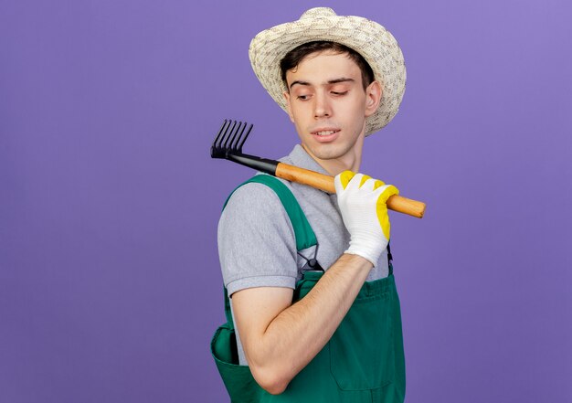 Fiducioso giovane giardiniere maschio che indossa cappello e guanti da giardinaggio che tengono il rastrello sulla spalla