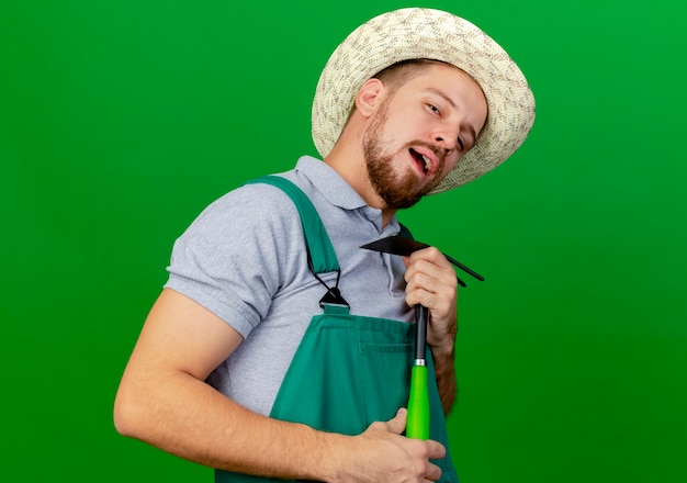 Fiducioso giovane giardiniere slavo bello in uniforme e cappello che tiene zappa guardando fingere di cantare usando zappa come microfono