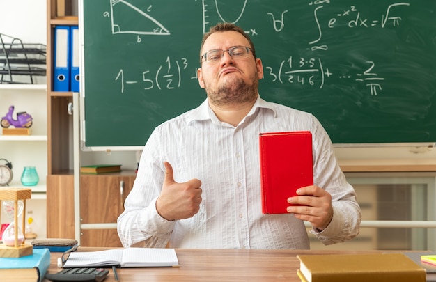 Fiducioso giovane insegnante con gli occhiali seduto alla scrivania con forniture scolastiche in aula tenendo il libro chiuso che mostra il pollice in alto guardando davanti