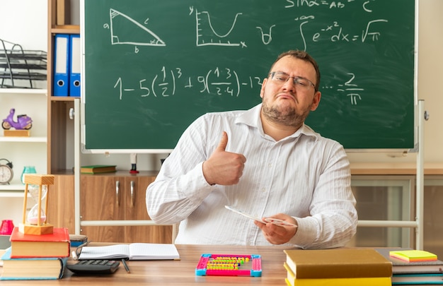 fiducioso giovane insegnante con gli occhiali seduto alla scrivania con materiale scolastico in aula tenendo il numero di fan guardando davanti mostrando pollice in su