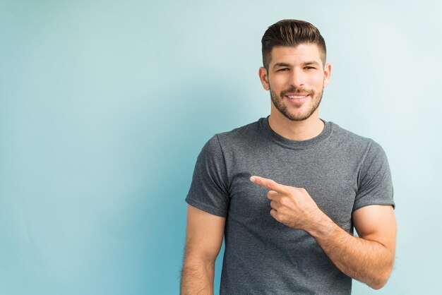 Fiducioso giovane latino che indossa una maglietta grigia e indica uno spazio vuoto mentre stabilisce il contatto visivo su uno sfondo semplice
