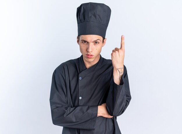 Fiducioso giovane maschio biondo cuoco in uniforme da chef e berretto che guarda l'obbiettivo che guarda l'obbiettivo rivolto verso l'alto isolato sul muro bianco con spazio di copia