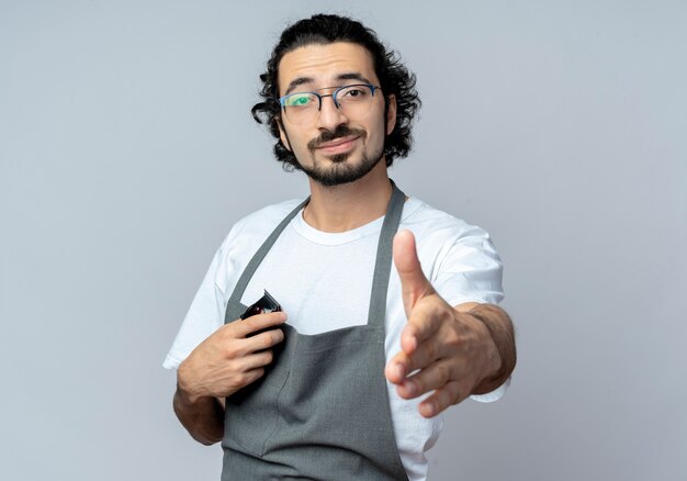 Fiducioso giovane maschio caucasico barbiere con gli occhiali e fascia per capelli ondulati in uniforme che tiene tagliatrici di capelli che allunga la mano alla macchina fotografica che gesturing ciao isolato su fondo bianco con lo spazio della copia