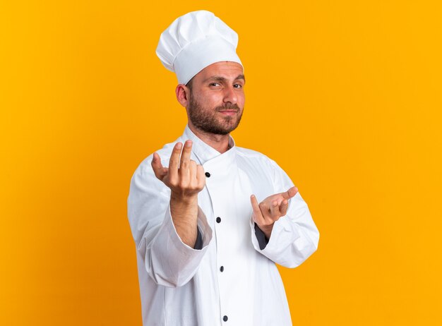 Fiducioso giovane maschio caucasico cuoco in uniforme da chef e berretto guardando la telecamera facendo venire qui gesto isolato sulla parete arancione