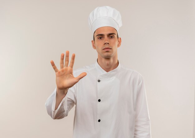 fiducioso giovane maschio cuoco che indossa l'uniforme dello chef che mostra il gesto di arresto sul muro bianco isolato